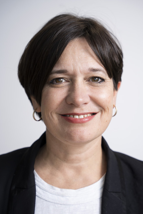 A head and shoulders shot of Jo Shanmugalingam, who has short dark hair and is wearing a white t shirt, black blazer and small gold hoop earrings