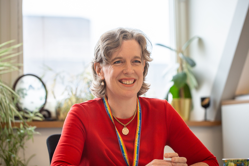 Emily Miles, a white woman with curly hair, sits with her elbows on a desk in an office. She is wearing a bright red dress, a rainbow lanyard and a gold pendant and is smiling. There is a window with plants on the windowsill behind her.
