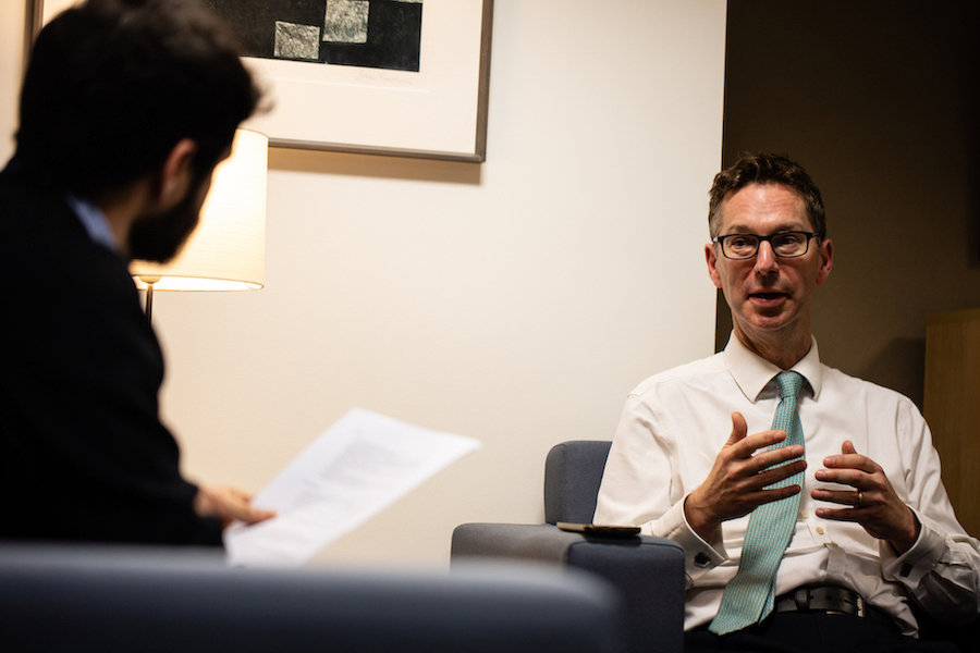 Schofield sits in an armchair in an office, talking. Across from him is an interviewer with dark hair and a beard, holding an A4 sheet of paper