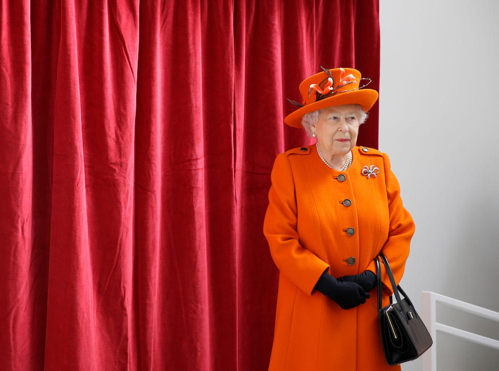 The Queen, in her later years with white hair, wearing an orange coat and hat, stands in front of a red curtain